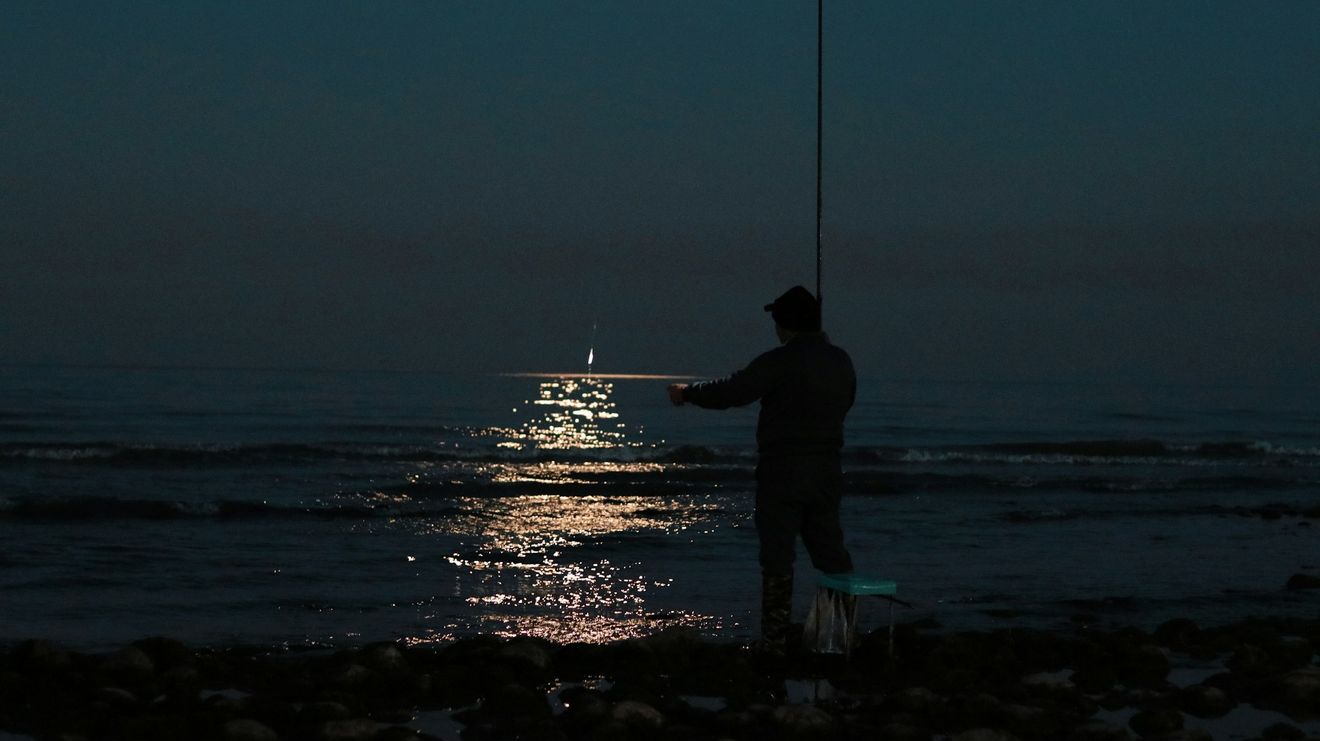 Un pescatore di notte in riva al mare, con la luna che si riflette sull’acqua.